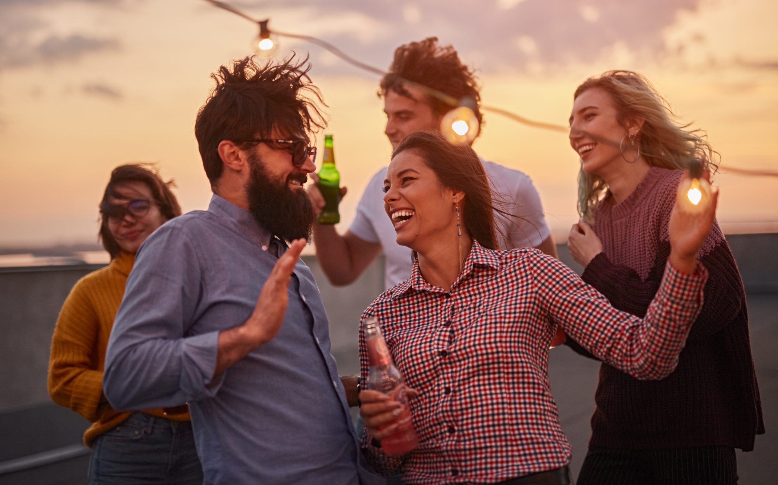 Group of joyful young friends in casual clothes drinking beer and dancing while enjoying summer party on rooftop at sunset time