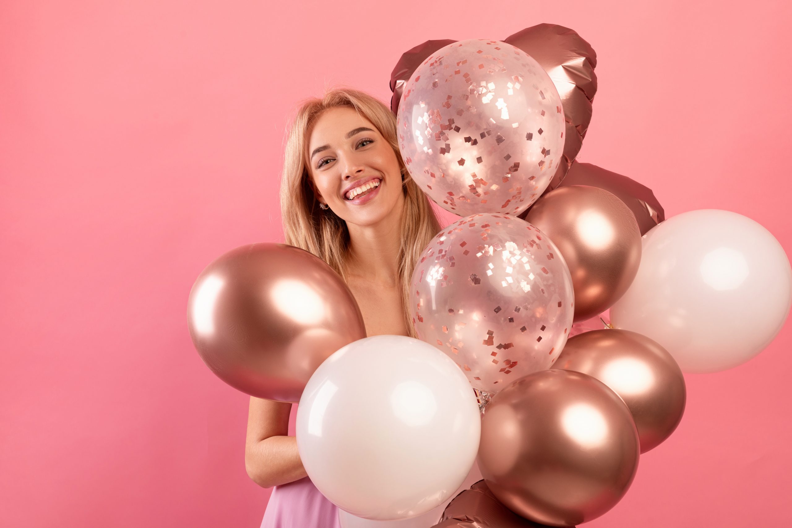 Charming millennial woman with bunch of air balloons having holiday party over pink studio background. Cheerful blonde having fun on her birthday celebration. Festive concept